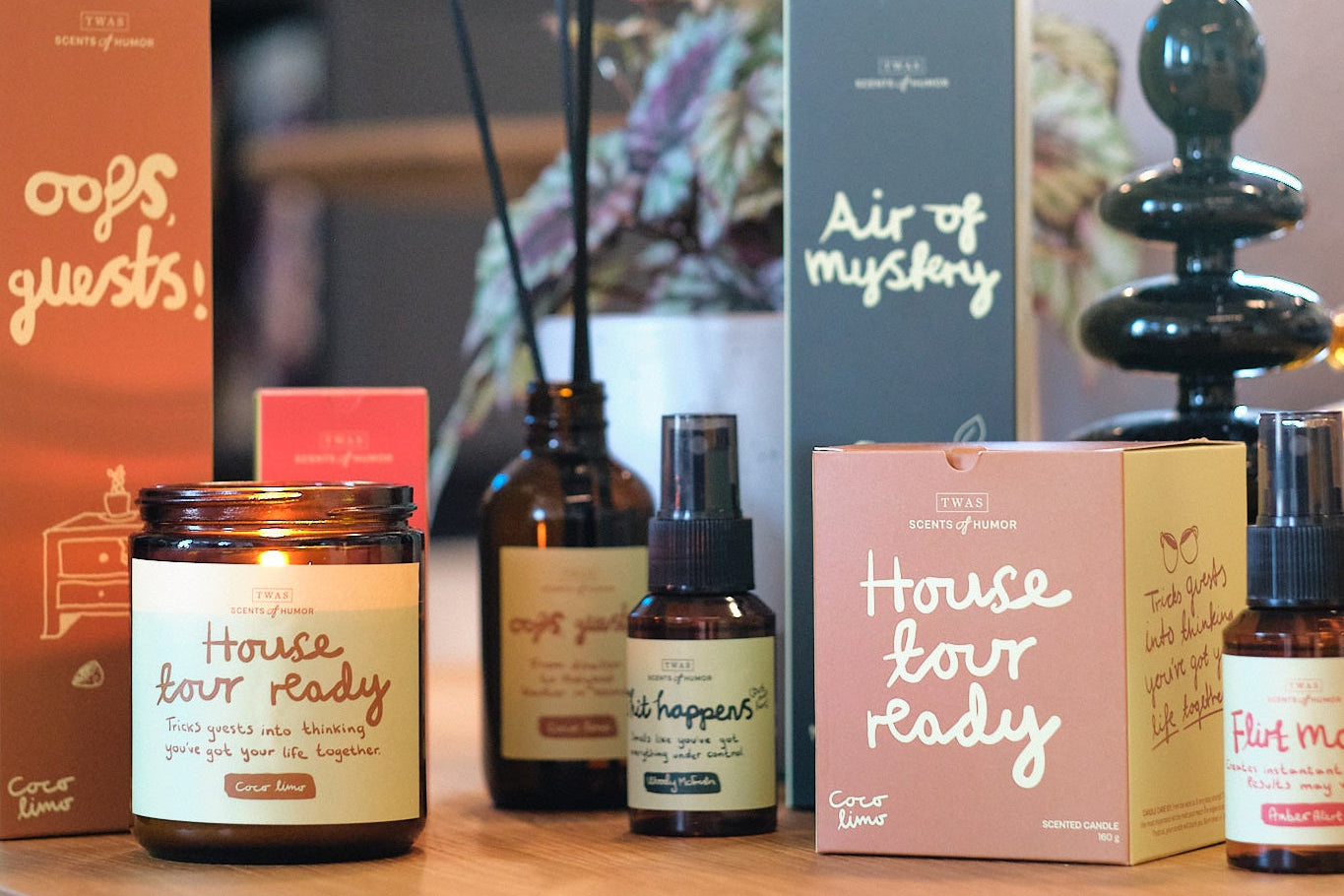 Scents of Humor Romduftserie fra TWAS, duftpinner, duftlys og romspray på et bord med en plante og lysestake